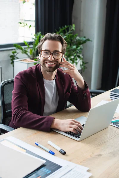 Empresário sorridente falando no smartphone e usando laptop no escritório — Fotografia de Stock