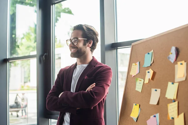 Smiling businessman with crossed arms standing near board with sticky notes in office — Stock Photo