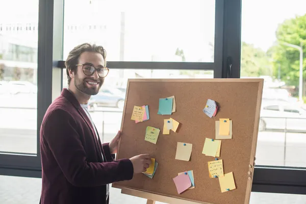 Hombre de negocios positivo en gafas de vista mirando a la cámara cerca del tablero con notas adhesivas - foto de stock