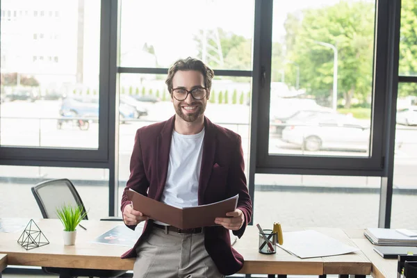 Glücklicher Geschäftsmann hält Papiermappe in der Hand und blickt in die Kamera am Arbeitstisch — Stockfoto