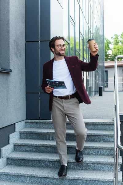 Cheerful businessman with paper cup and document walking on stairs near building — Stock Photo