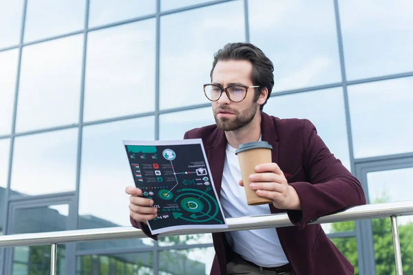 Businessman looking at document and holding coffee to go near railing and building outdoors — Stock Photo