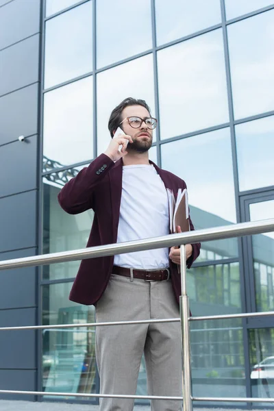 Jeune homme d'affaires en tenue formelle parlant sur smartphone et tenant des dossiers en papier à l'extérieur — Photo de stock