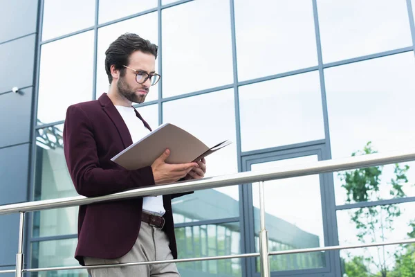 Бизнесмен в очках смотрит на бумажную папку возле здания на открытом воздухе — стоковое фото