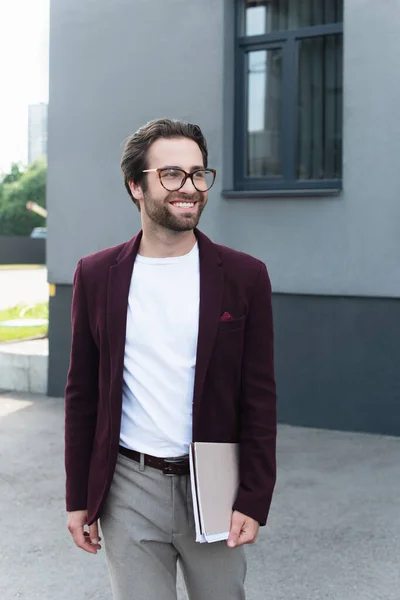 Cheerful businessman in eyeglasses holding paper folders near building outdoors — Stock Photo