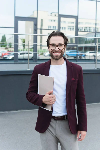 Homme d'affaires dans l'usure formelle tenant dossiers en papier et souriant à la caméra à l'extérieur — Photo de stock