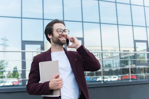 Uomo d'affari positivo con cartelle di carta che parla su smartphone sulla strada urbana — Foto stock