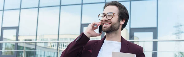 Junger Geschäftsmann mit Papiermappe lächelt, während er draußen auf dem Smartphone spricht, Banner — Stockfoto