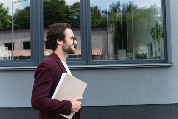 Vue latérale d'un homme d'affaires souriant avec des chemises en papier marchant près du bâtiment à l'extérieur — Photo de stock