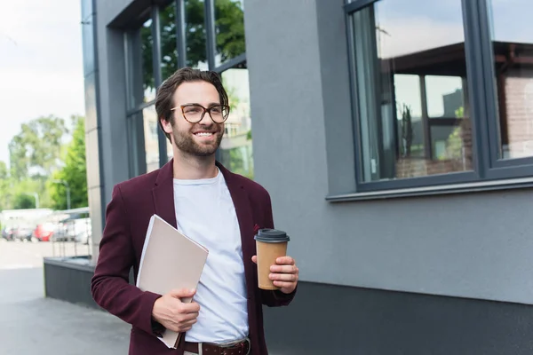 Cheerful businessman with coffee to go and paper folder walking on urban street — Stock Photo