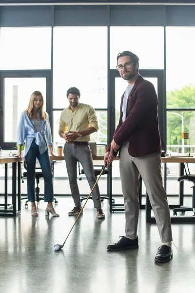 Молодой бизнесмен играет в гольф рядом с размытыми коллегами в офисе — стоковое фото