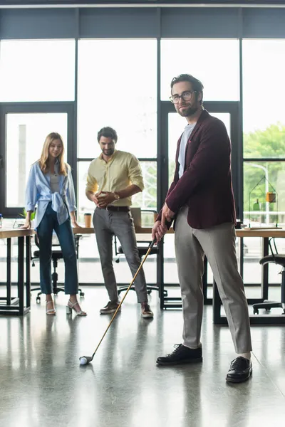 Junger Geschäftsmann spielt Golf in der Nähe verschwommener Kollegen im Büro — Stockfoto