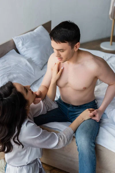 High angle view of sensual young woman in silk robe touching chest of smiling boyfriend in bedroom — Stock Photo