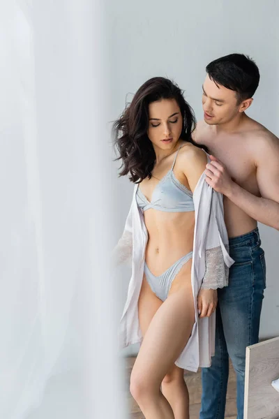 Hombre apasionado en jeans quitándose la bata de seda en la mujer sensual en ropa interior sexy - foto de stock