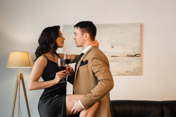 Vista lateral del hombre levantando pierna de mujer seductora en vestido de deslizamiento con copa de vino tinto - foto de stock