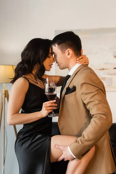 Vue latérale de l'homme passionné levant jambe de femme séduisante en robe glissante avec verre de vin rouge — Photo de stock