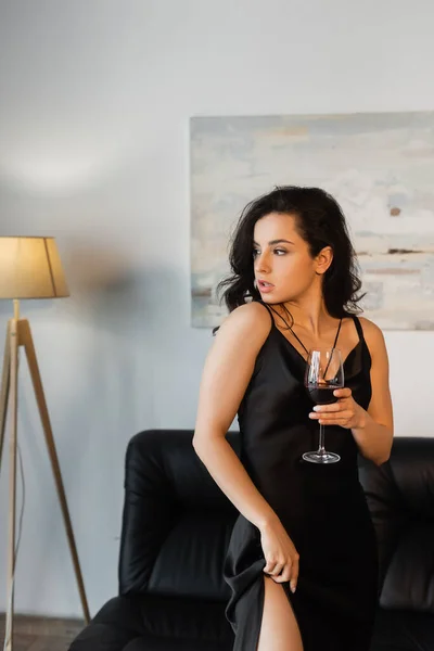 Séduisante femme en robe noire glissante tenant un verre de vin et assise sur un canapé — Photo de stock