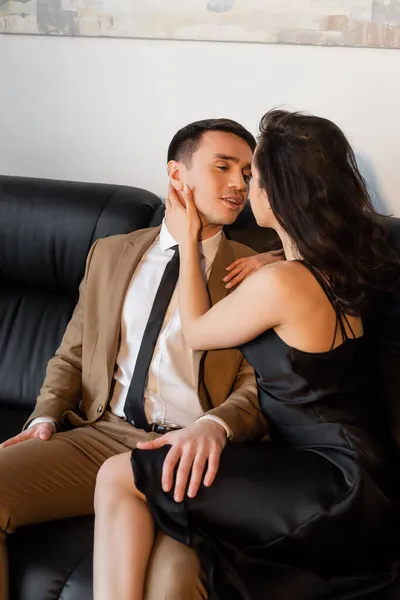 Brünette Frau im schwarzen Slip-Kleid berührt Gesicht eines Mannes auf Sofa — Stockfoto