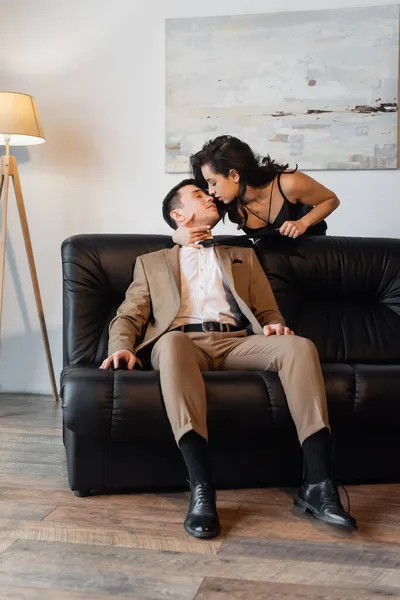 Mulher morena no vestido de deslizamento puxando gravata enquanto seduz o homem no sofá — Fotografia de Stock