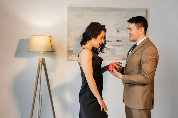 Vista lateral do homem feliz segurando caixa de presente em forma de coração perto namorada surpreso em vestido de deslizamento preto — Fotografia de Stock