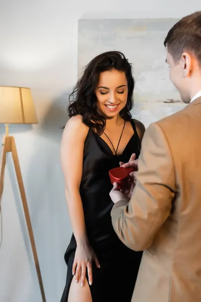 Verschwommener Mann mit herzförmiger Geschenkschachtel neben lächelnder Freundin im schwarzen Slip-Kleid — Stockfoto