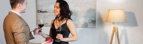 Hombre feliz sosteniendo en forma de corazón caja de regalo y vaso de vino cerca de novia sonriente en vestido de deslizamiento negro, bandera - foto de stock