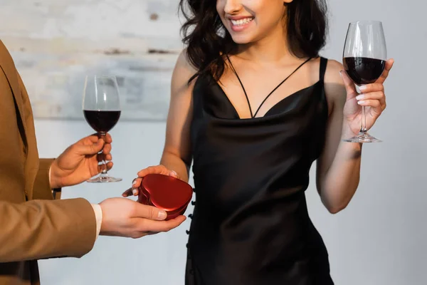 Обрезанный вид человека, держащего в руках подарочную коробку в форме сердца и бокал вина рядом с счастливой девушкой в черном платье скольжения — стоковое фото