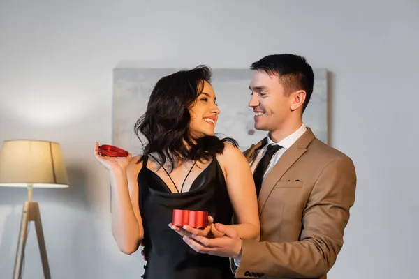 Mulher feliz em vestido de deslizamento preto segurando caixa de presente em forma de coração enquanto olha para o namorado — Fotografia de Stock