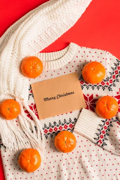 Top view of greeting card with merry christmas lettering near tangerines on knitted sweater isolated on red — Stock Photo
