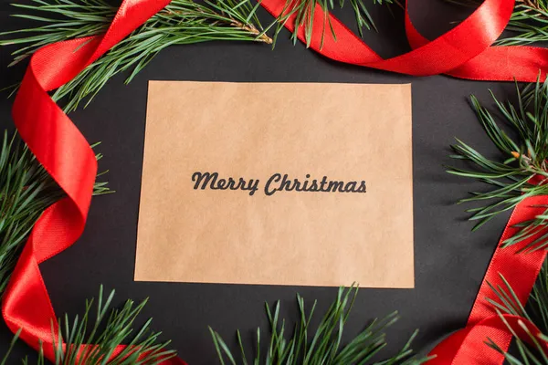Top view of greeting card with merry christmas lettering near fir branches and red ribbon on black — Stock Photo