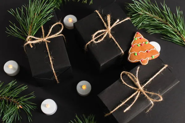 Top view of gingerbread cookie on wrapped dark gift boxes near candles and pine branches on black — Stock Photo