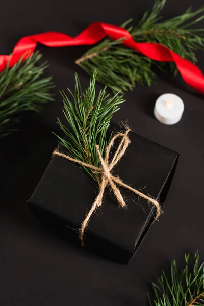 Top view of gift box with fir branch near candle and blurred red ribbon on black — Stock Photo