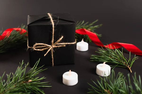 Wrapped present near fir branches and candles with blurred red ribbon on black background — Stock Photo