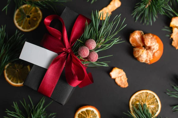 Vista superior de la caja de regalo envuelta con cinta roja y tarjeta en blanco cerca de mandarinas borrosas en negro - foto de stock