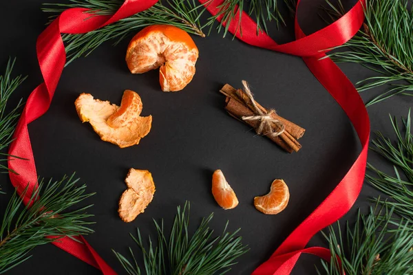 Top view of peeled tangerines and cinnamon sticks near red ribbon and fir branches on black — Stock Photo