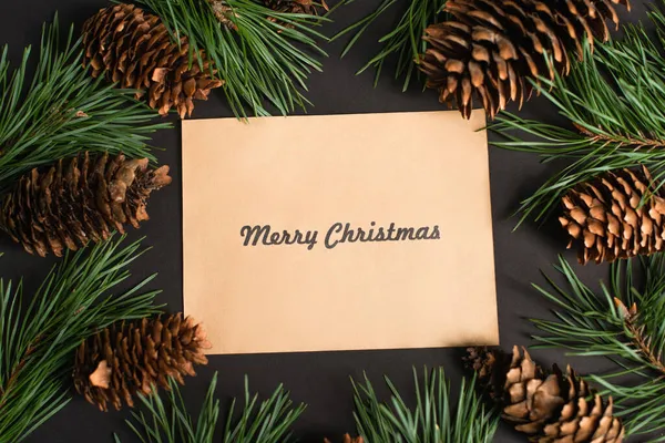 Top view of craft paper with merry christmas lettering near fir branches and pine cones on black — Stock Photo