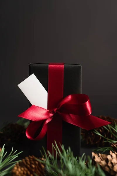 Caja de regalo envuelta con cinta roja y tarjeta en blanco cerca de ramas de abeto borrosas y conos de pino en negro - foto de stock