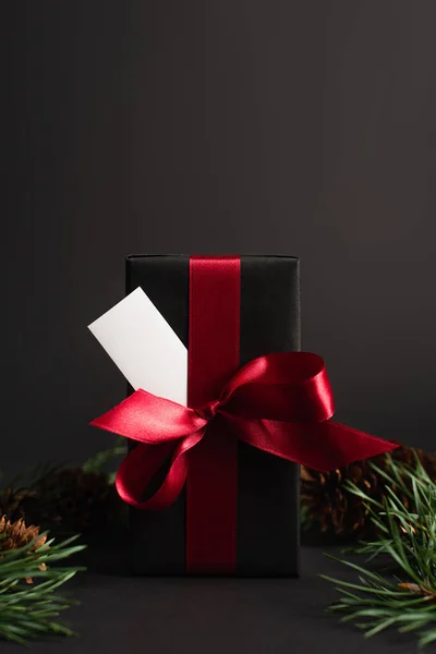 Wrapped present with red ribbon and blank card near blurred fir branches on black — Stock Photo