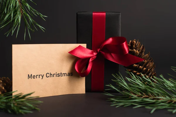 Papel artesanal con letras de navidad feliz cerca de caja de regalo y ramas de abeto en negro — Stock Photo