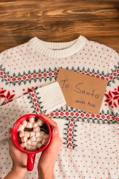 Cropped view of woman holding cocoa with marshmallows near blurred letter to santa near on knitted sweater — Stock Photo