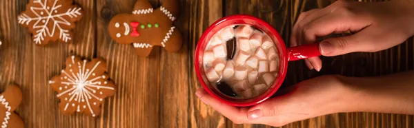Cropped view of woman holding cup of cocoa with marshmallows near gingerbread cookies on wooden surface, banner — Stock Photo
