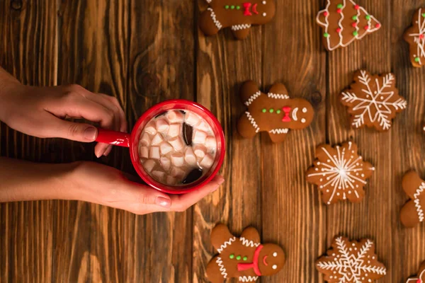 Cropped view of woman holding cup of cocoa with marshmallows near gingerbread cookies on wooden surface — Stock Photo