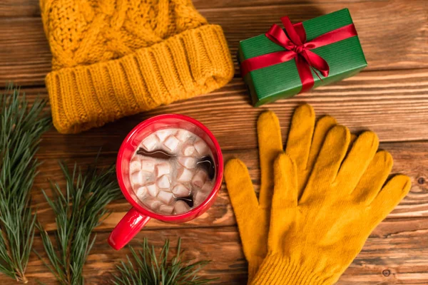 Vue du dessus du chapeau et des gants tricotés jaunes près de la boîte cadeau, tasse de cacao avec guimauves et branches sur la surface en bois — Photo de stock