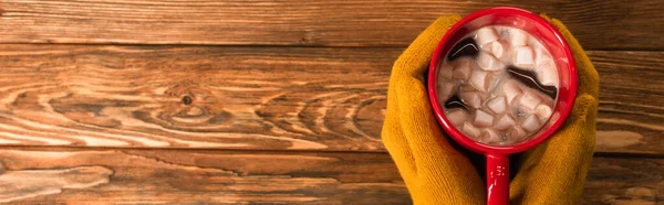 Vista recortada de la persona en guantes amarillos sosteniendo taza de cacao con malvaviscos sobre la superficie de madera, pancarta - foto de stock