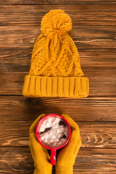 Cropped view of person in yellow gloves holding cup of cocoa with marshmallows near hat on wooden surface — Stock Photo