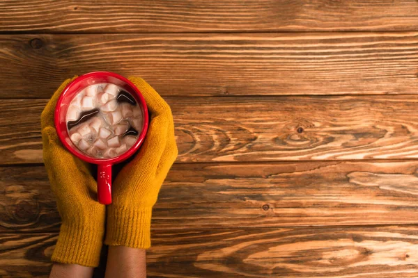 Cropped view of person in yellow gloves holding cup of cocoa with marshmallows above wooden surface — Stock Photo