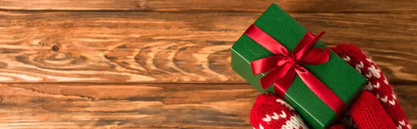 Cropped view of person in red mittens holding wrapped gift box above wooden surface, banner — Stock Photo