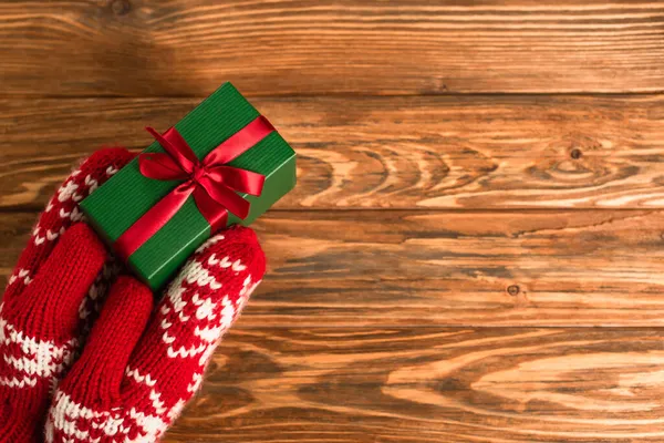 Cropped view of person in red mittens holding wrapped gift box above wooden surface — Stock Photo