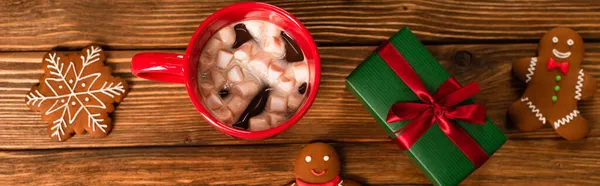 Top view of cup of cocoa with marshmallows near gift box and gingerbread cookies on wooden surface, banner — Stock Photo