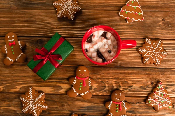 Top view of cup of cocoa with marshmallows near gift box and gingerbread cookies on wooden surface — Stock Photo